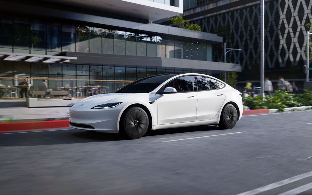 White Tesla Model 3 driving past a glass office building