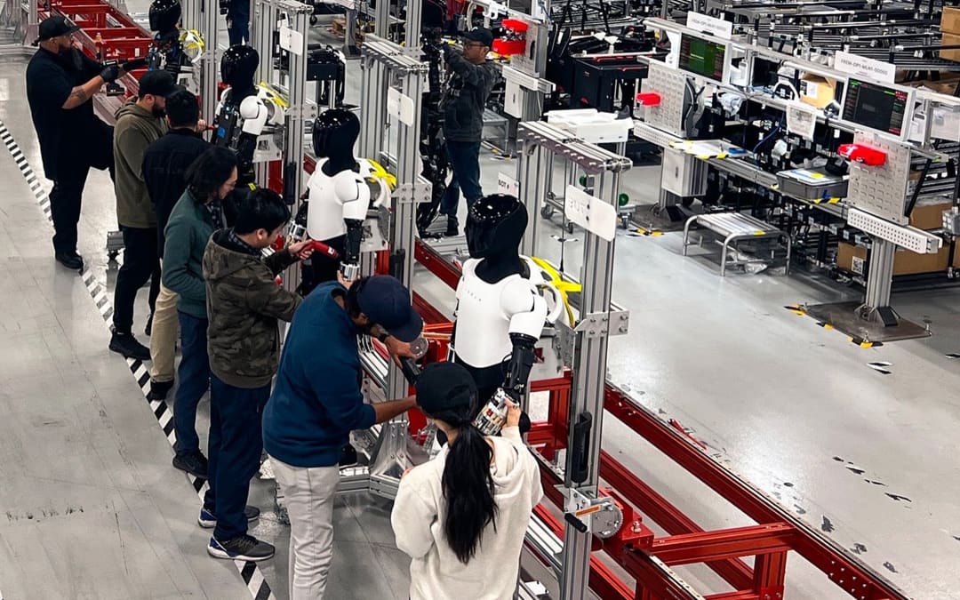Optimus robots on a factory line with Tesla staff working on them (@Tesla_Optimus “Me, Robot” post)